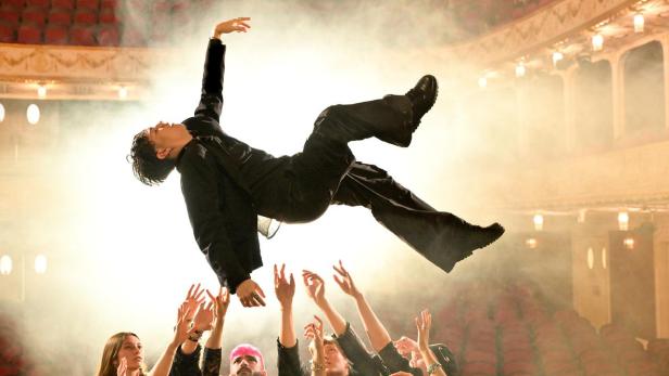 Ein Mann in schwarzer Kleidung wird von einer Menschenmenge auf einer Bühne in die Luft gehoben.