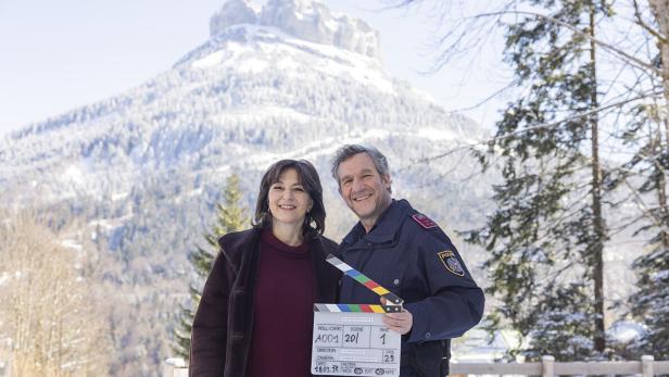 Ein Mann in Polizeiuniform und eine Frau halten eine Filmklappe vor einer verschneiten Bergkulisse.
