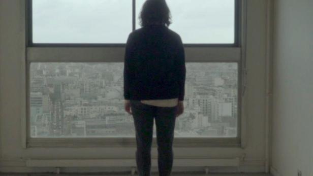 Eine Person steht in einem leeren Raum und blickt aus dem Fenster auf die Stadt.