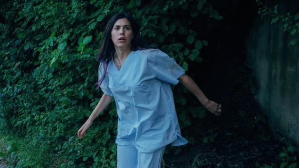 Eine Frau in Arztkleidung wirkt erschrocken, während sie durch ein Gebüsch läuft.
