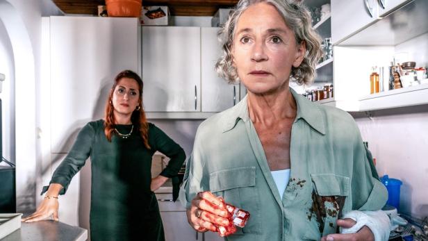 Eine ältere Frau mit blutverschmierter Hand und Gipsverband steht besorgt in einer Küche, während eine jüngere Frau im Hintergrund steht.