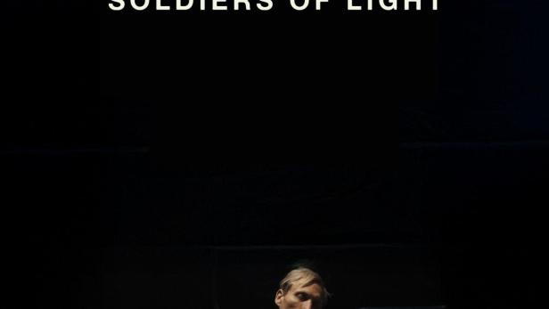 Ein Mann sitzt auf einem Stuhl vor einem schwarzen Hintergrund mit dem Text „Soldiers of Light“.