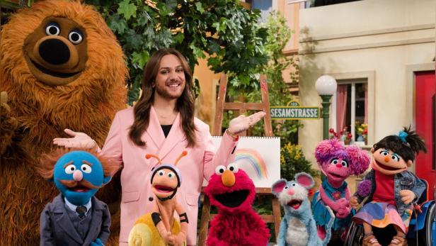 Riccardo Simonetti mit verschiedenen Puppen aus der Sesamstraße vor einem gemalten Regenbogen.