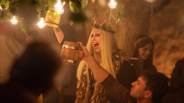 Eine Frau mit Geweihkrone und Fellschal prostet mit einem Holzkrug in die Menge.