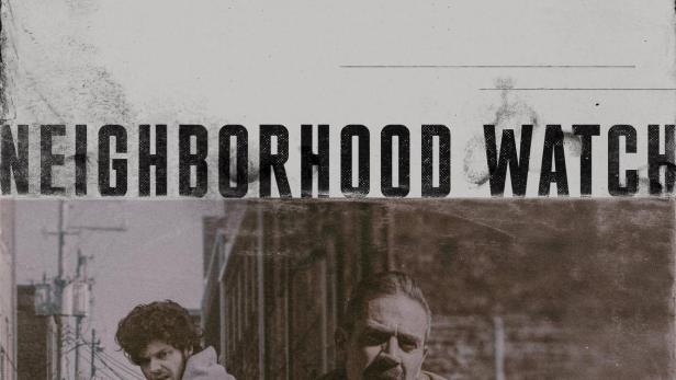 Ein Mann mit Revolver lehnt an einem Auto, während ein anderer Mann im Hintergrund steht.