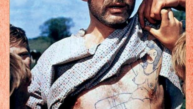 Ein Mann mit Hut und einem Tattoo auf der Brust, Szene aus „Die Heiden von Kummerow“.
