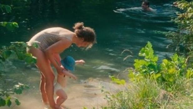 Eine Frau mit Kind am Ufer eines Sees, im Hintergrund schwimmt eine Person.