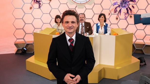 Ein Mann im Anzug steht vor einem Fernsehstudio-Set mit drei Personen im Hintergrund.