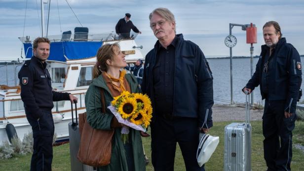 Eine Frau mit Sonnenblumenstrauß und Polizisten am Hafen, möglicherweise eine Szene aus „Morden auf Ödinsel“.
