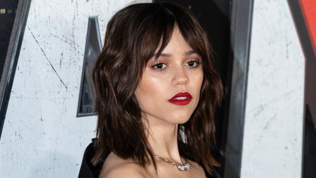 Ein Porträt von Jenna Ortega mit roten Lippen und braunem, gewelltem Haar.