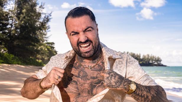 Cosimo Citiolo posiert am Strand und reißt sein Hemd auf, um seine Tätowierungen zu zeigen.