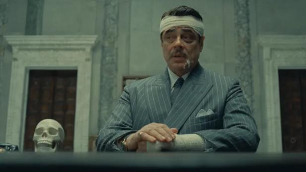 Ein Mann mit Verbänden am Kopf und Arm sitzt an einem Tisch mit einem Totenkopf.