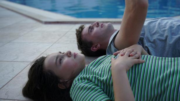 Ein Mädchen und ein Mann liegen auf Fliesen neben einem Pool und schauen nach oben.