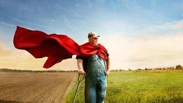 Ein Landwirt mit rotem Umhang und Spaten steht auf einem Feld.