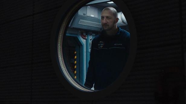 Ein Mann in Astronautenkleidung schaut aus einem Bullauge in dem Film „U Are the Universe“.