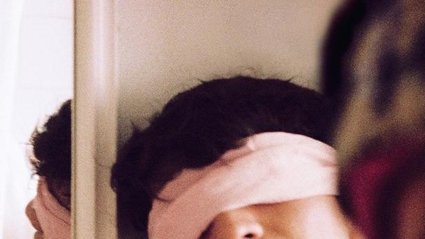 Eine Person mit einer rosa Augenbinde steht vor einem Spiegel.