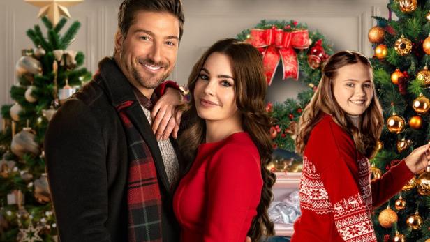 Ein Mann und eine Frau umarmen sich vor einem Weihnachtsbaum, während ein Mädchen ihn schmückt.