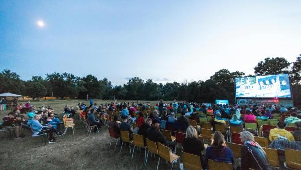 Besucher sitzen auf Stühlen im grünen vor einer Leinwand, ein Open-Air-Kino. 