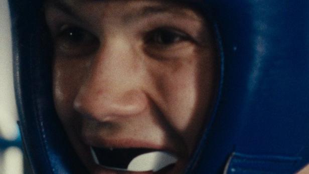 Ein Boxer mit blauem Kopfschutz und Mundschutz streckt die Zunge heraus.