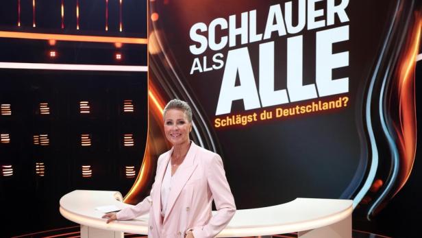 Moderatorin steht vor dem Logo der Quizsendung „Schlauer als alle“.
