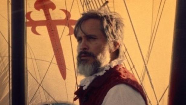 Ein Mann mit Bart und Weste steht auf einem Segelschiff mit dem Santiago-Kreuz.