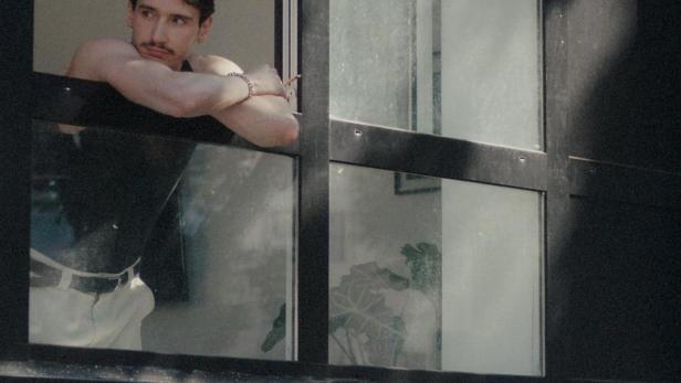 Ein Mann mit Schnurrbart lehnt am Fenster und schaut nach draußen.