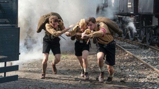 Drei uniformierte Männer mit Gepäck fliehen vor einer Explosion in der Nähe eines Zuges.