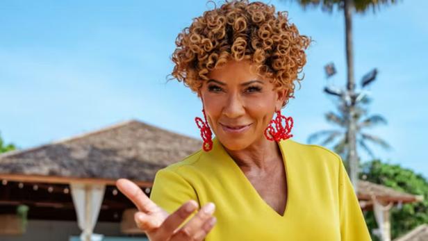 Eine Frau mit lockigem Haar und roten Ohrringen vor einem Strand-Resort.