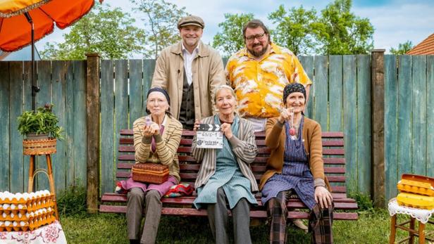 Drei ältere Damen sitzen mit zwei Männern und einer Filmklappe für „Eierkratz-Komplott“ auf einer Bank.