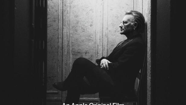 Schwarzweiß-Porträt von Bono, dem Sänger von U2, im Profil vor einer Wand sitzend.