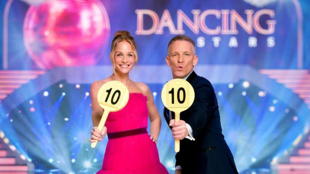 Eine Frau in einem pinkfarbenen Kleid und ein Mann im Anzug halten Wertungstafeln mit der Zahl 10 vor dem „Dancing Stars“-Hintergrund.