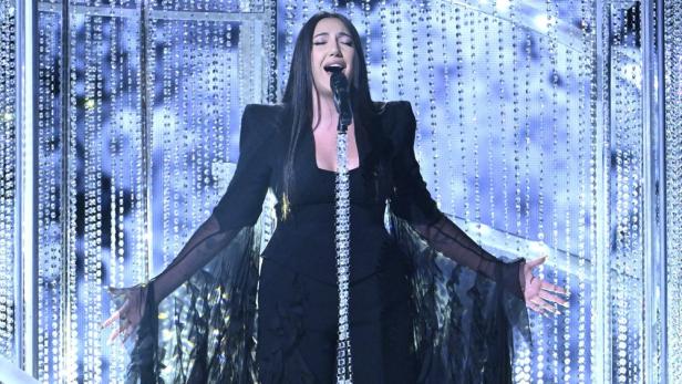 Eine Sängerin mit dunklem Haar und einem schwarzen Kleid singt auf einer Bühne mit Kristallvorhängen.