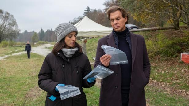 Zwei Ermittler betrachten Beweismittel in Plastiktüten an einem Tatort im Freien.