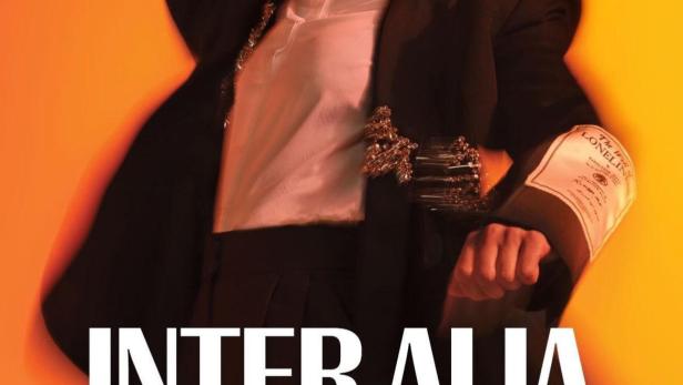 Rosamund Pike posiert in einem schwarzen Hosenanzug vor einem orangefarbenen Hintergrund.