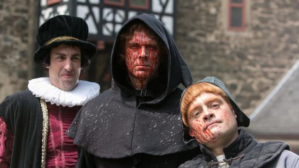 Drei Schauspieler in historischen Kostümen und mit Blut im Gesicht stehen vor einer Burg.