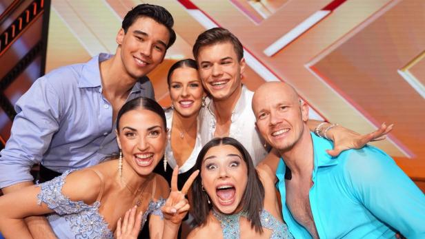 Eine Gruppe von Tänzern posiert lächelnd vor einem X-förmigen Hintergrund.