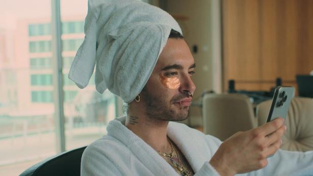 Bill Kaulitz mit Handtuch auf dem Kopf und Augenpads, während er auf sein Handy schaut.