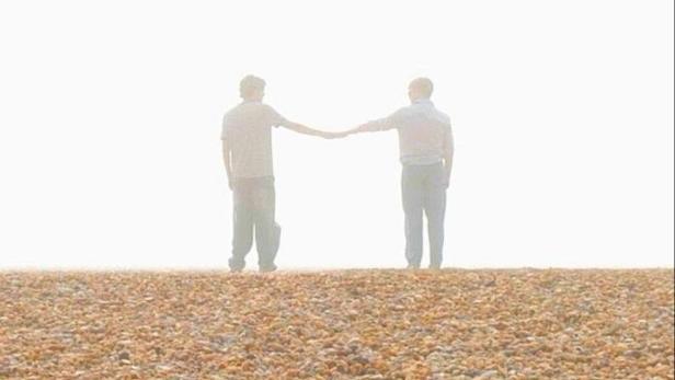Das Filmplakat für „Him“ zeigt zwei Männer, die sich an einem Strand die Hände reichen.