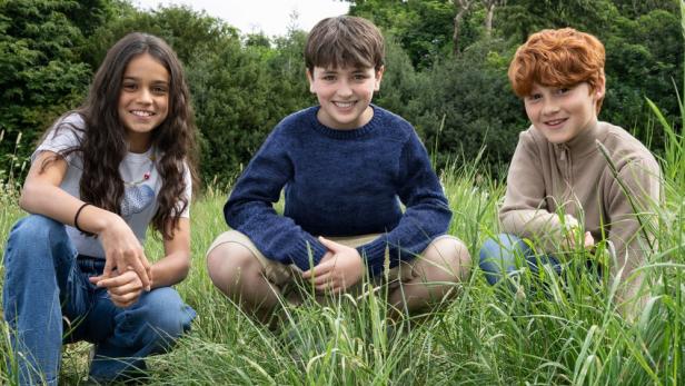 Drei lächelnde Kinder kauern im hohen Gras, möglicherweise Darsteller einer „Harry Potter“-Serie.