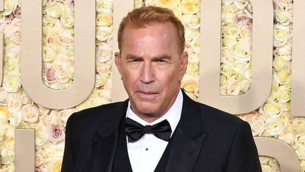 Kevin Costner in einem schwarzen Smoking vor einer Wand aus Rosen.