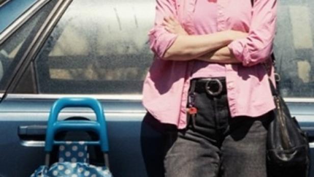 Eine Frau mit Sonnenbrille und rosa Hemd steht neben einem blauen Auto.