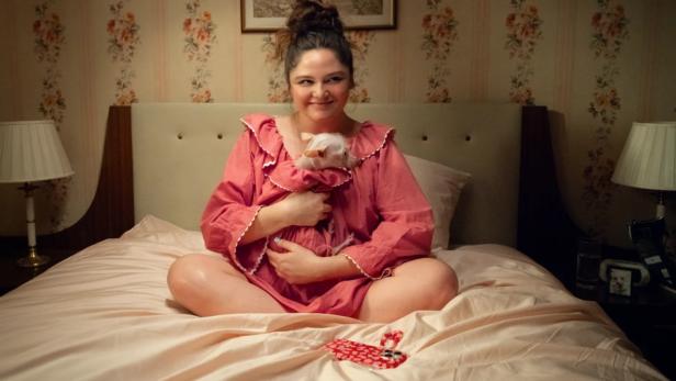 Eine Frau sitzt im Schneidersitz auf einem Bett und hält eine kleine, weiße Stoffpuppe im Arm.