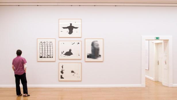 Ein Mann betrachtet in einer Galerie eine Wand mit fünf abstrakten, gerahmten Bildern.