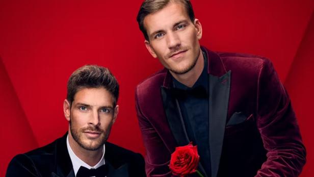 Zwei Männer im Anzug posieren vor einem roten Hintergrund, einer hält eine rote Rose.