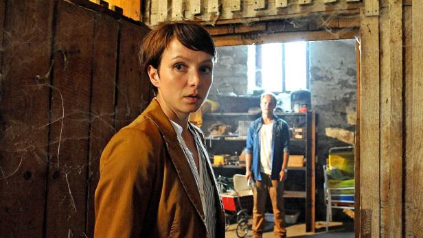 Eine Frau blickt besorgt in die Kamera, während ein Mann im Hintergrund steht.