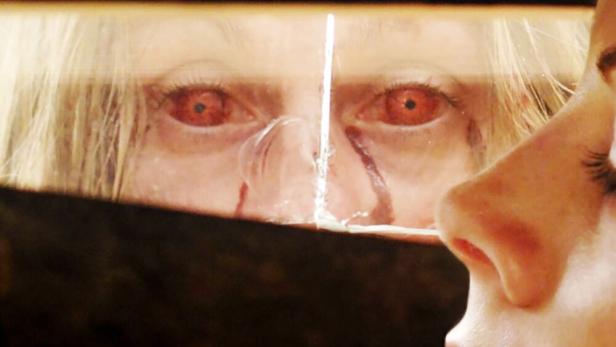 Eine Frau blickt in einen Spiegel, in dem eine andere Frau mit roten Augen und Blut im Gesicht zu sehen ist.