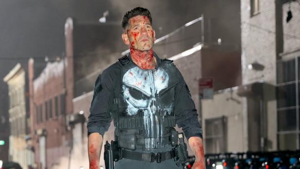 Jon Bernthal als der Punisher, blutverschmiert und in Kampfausrüstung, in einer dunklen Stadt.
