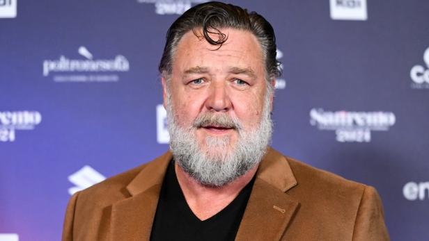 Porträt von Russell Crowe mit Bart und braunem Sakko vor einem blauen Hintergrund.