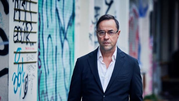 Ein Mann mit Brille und dunklem Anzug steht vor einer Wand mit Graffiti.