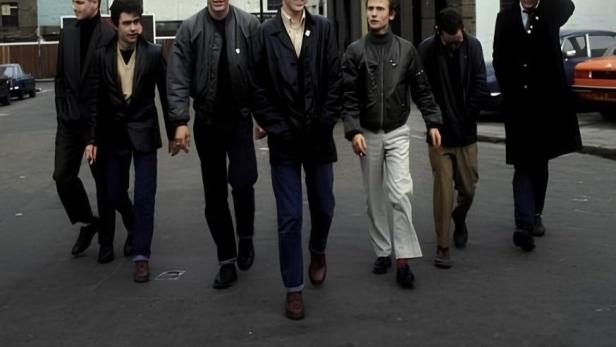 Die Band Madness geht eine Straße entlang, im Hintergrund Gebäude.